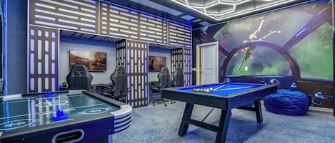 A pool table, two gaming chairs, a foosball table, and a large window with a view of a green landscape.