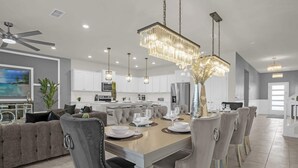 A modern kitchen with white cabinets, a central island, and a dining area with a wooden table and grey upholstered chairs. - Upstay: Family Retreat w Pool, Cinema & Games Room (Clermont)