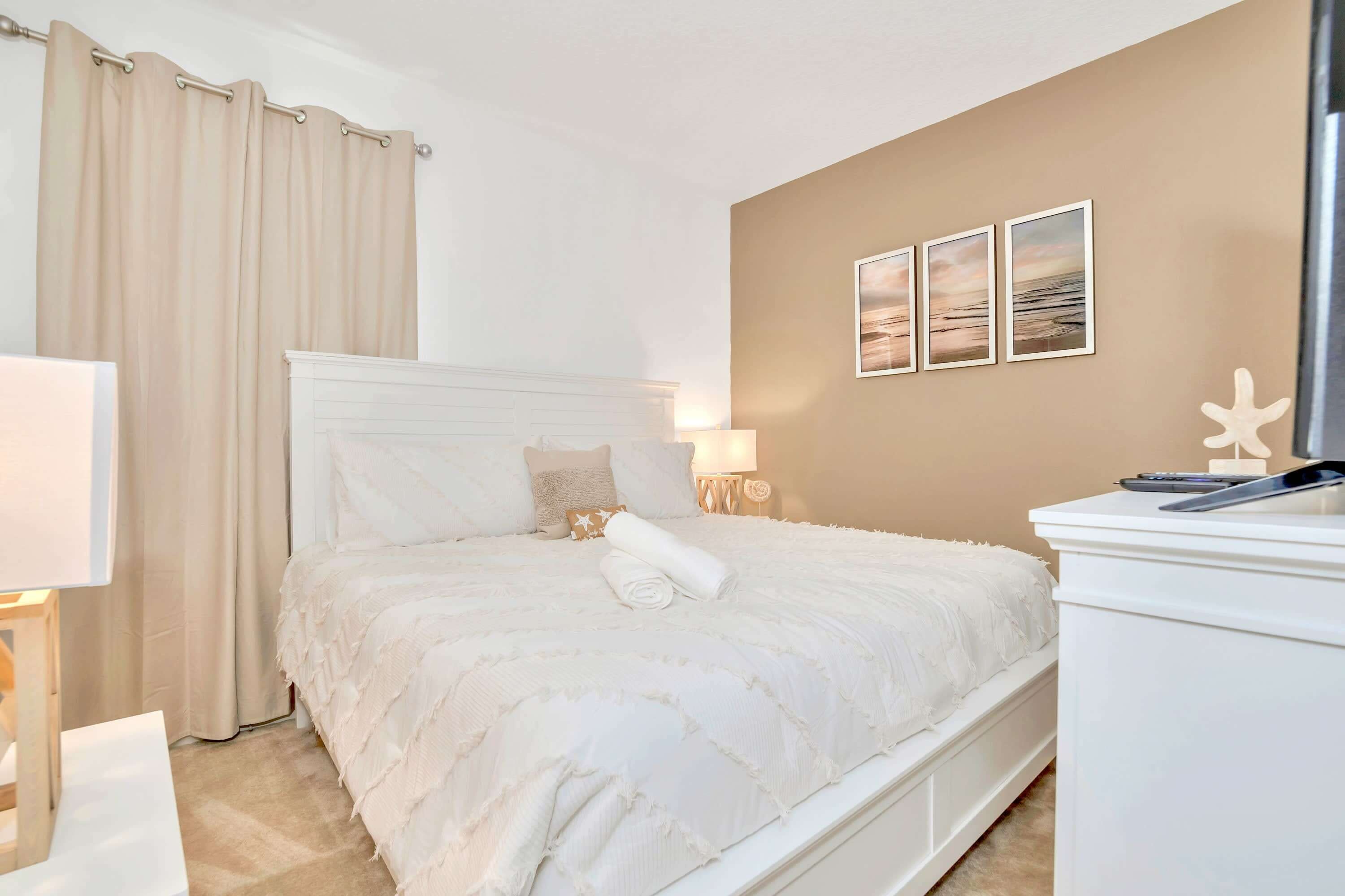A neatly made bed with a white headboard, a bedside table with a lamp, and a wall with three framed pictures.