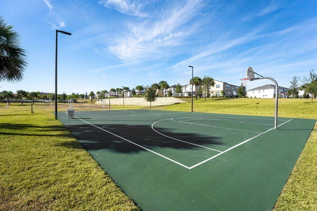 An outdoor basketball court with clear markings, surrounded by grass and trees, under a clear sky.