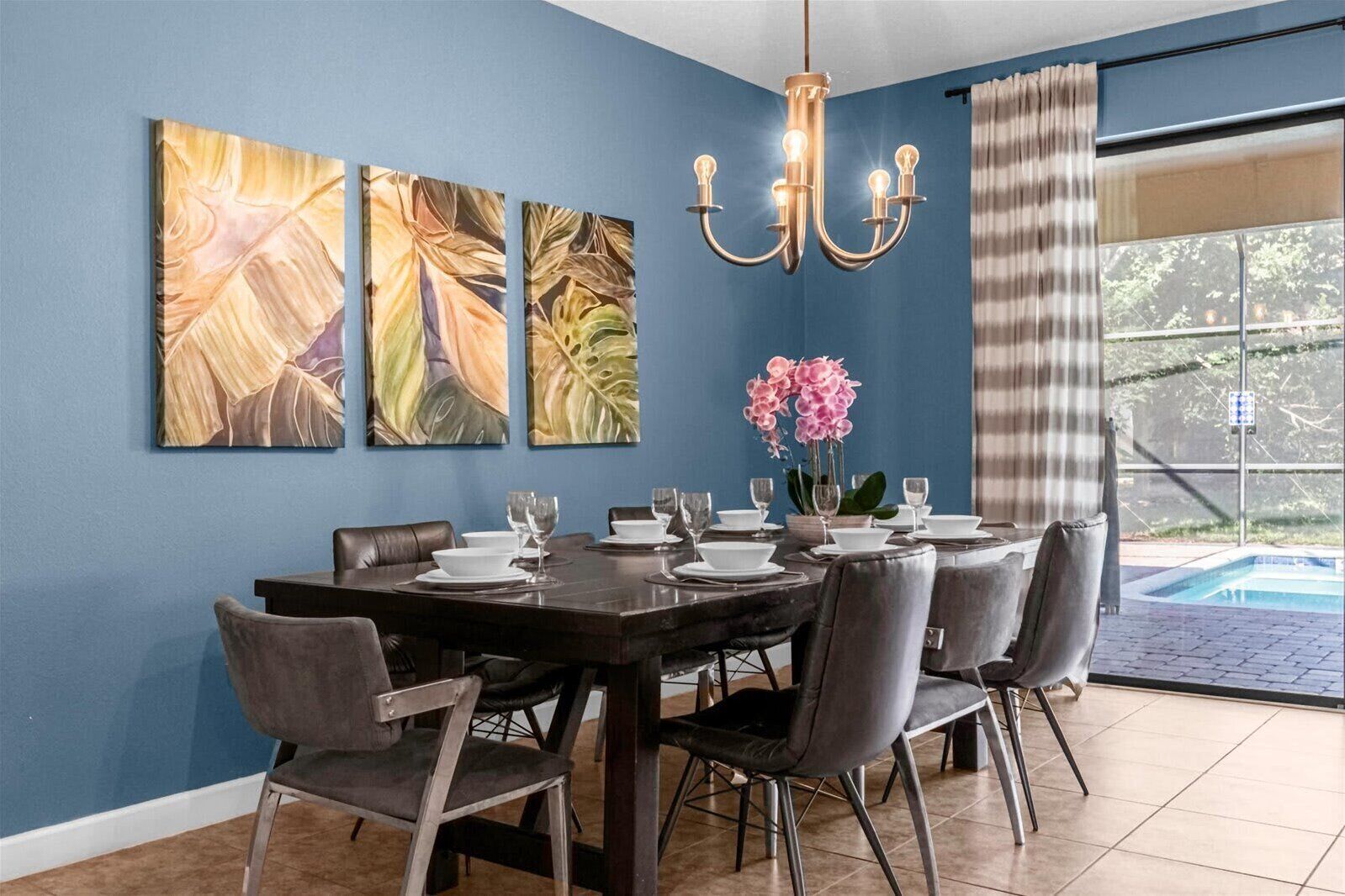 A dining area with a dark wooden table set for six, surrounded by grey upholstered chairs. There are three large abstract paintings on the wall, a chandelier above the table, and a view of a swimming pool through a large window with curtains.