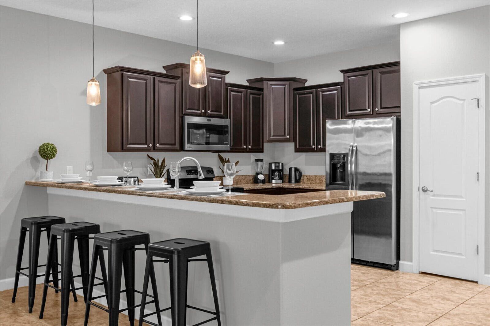 A modern kitchen with dark brown cabinets, a stainless steel refrigerator, a granite countertop, and black bar stools.