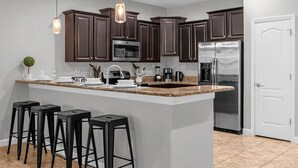 A modern kitchen with dark brown cabinets, a stainless steel refrigerator, a granite countertop, and black bar stools. - Upstay Pool Paradise Mansion Games Room & BBQ (Davenport)