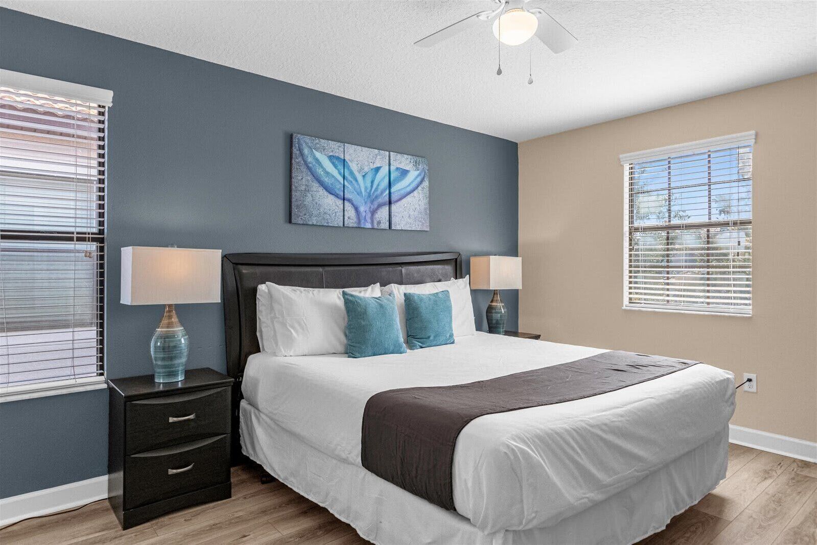 Un dormitorio con cabecera de madera oscura, ropa de cama blanca y almohadas azules decorativas. Hay una mesita de noche con una lámpara y una pequeña mesa con un florero. Se ve un ventilador de techo, y la habitación tiene una ventana con persianas.