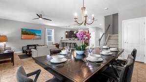 A spacious dining area with a long table set for six, a chandelier, and a view of the kitchen in the background. - Upstay Pool Paradise Mansion Games Room & BBQ (Davenport)
