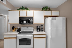 A compact kitchen with white appliances, including a stove, microwave, and refrigerator, with wooden accents and green plants on the countertop.