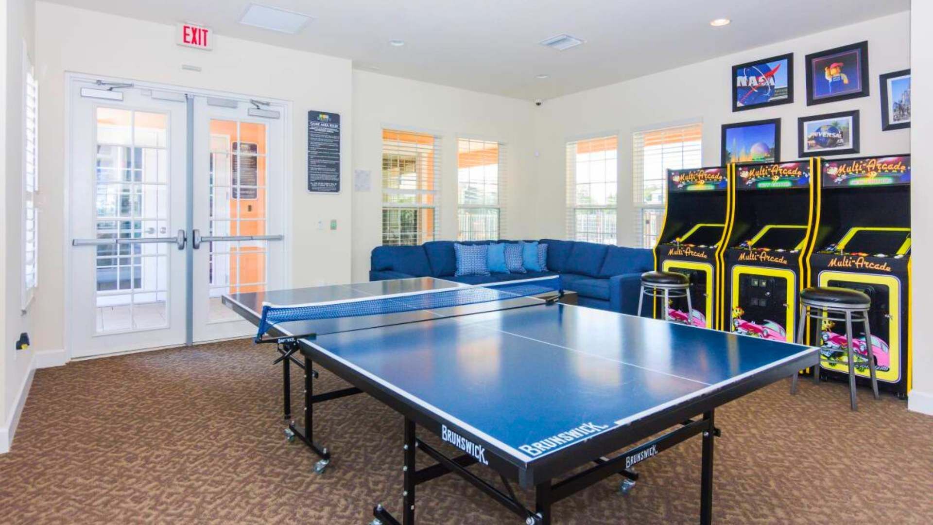 A table tennis table with a Brunswick logo, a blue sofa, and arcade machines labeled 'Multi-Arcade' in a room with carpeted flooring and white walls.