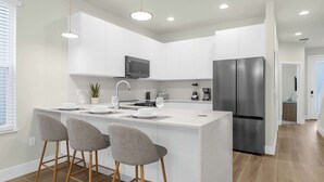 A modern kitchen with white cabinetry, a stainless steel refrigerator, a built-in microwave, and a central island with bar stools.