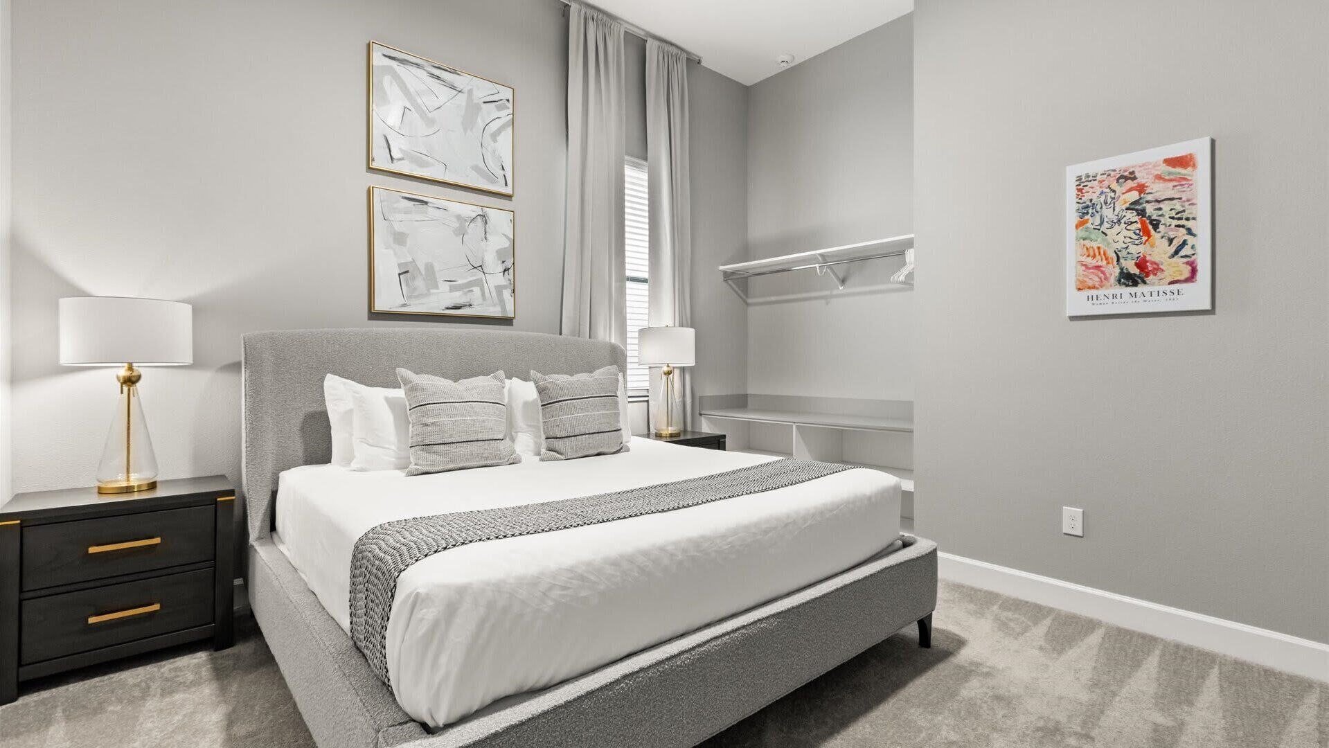 A modern bedroom with a grey bed, white bedding, and a grey carpet. There are two bedside tables with lamps, a large window with curtains, and a wall-mounted shelf.