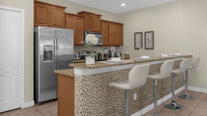 A modern kitchen with a stainless steel refrigerator, wooden cabinets, a granite countertop, and a bar area with bar stools. - Upstay: Pet-friendly Family Home w Pool & Sundeck (Kissimmee)
