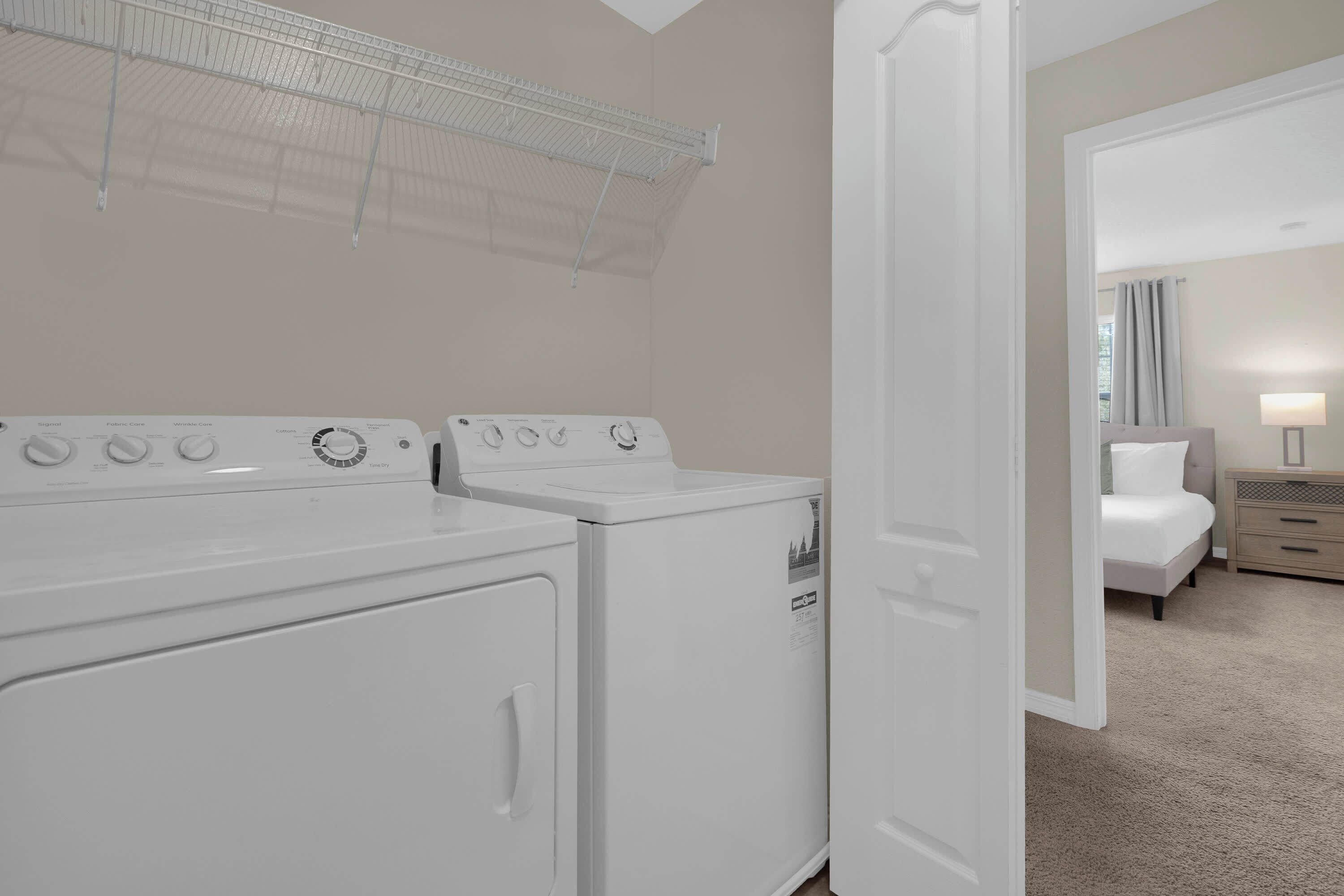 Une buanderie équipée de deux lave-linge et deux sèche-linge blancs, d’un étendoir métallique, et une chambre comprenant un lit, une table de chevet et une lampe.