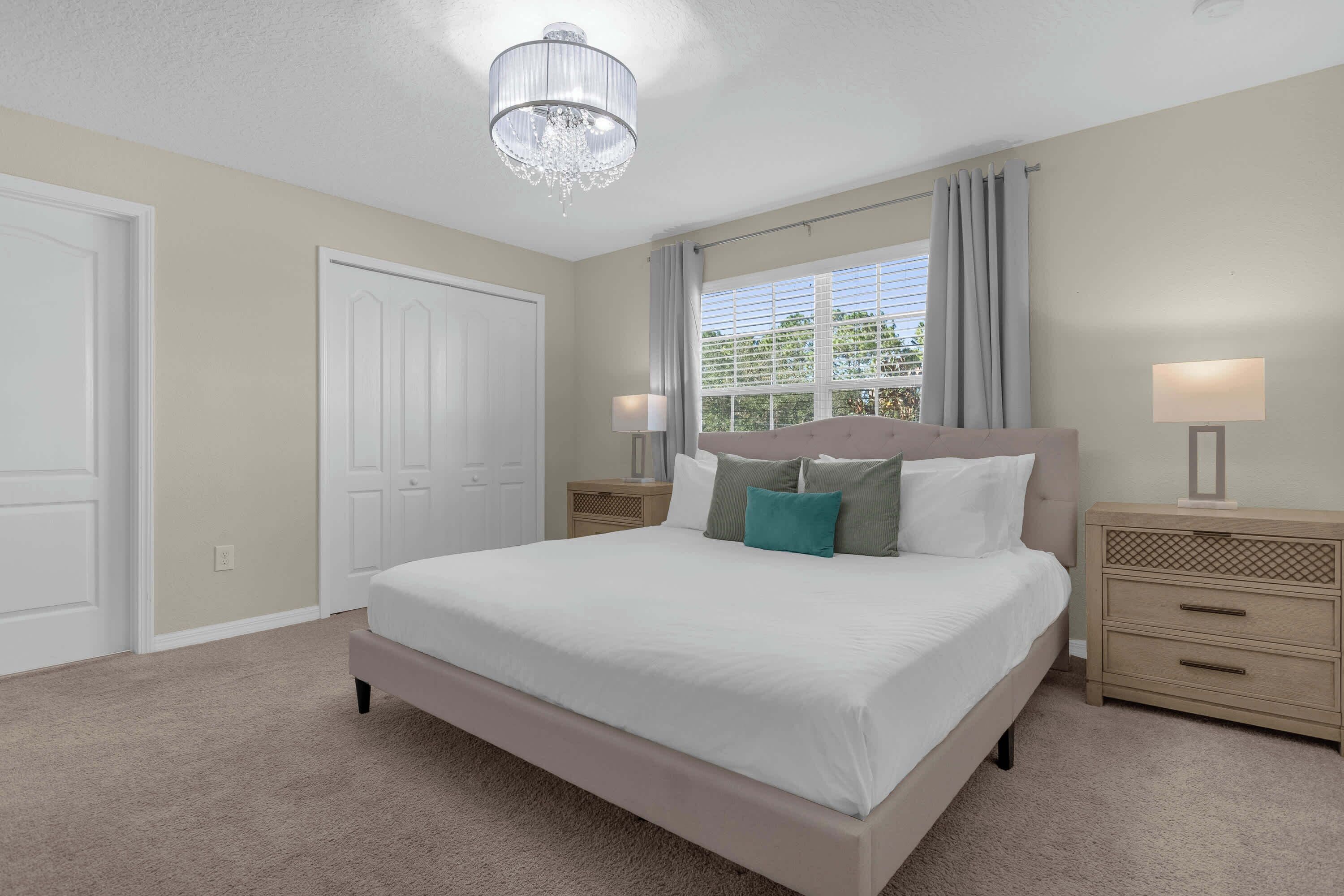 A bedroom with a large bed, two bedside tables, a chandelier, and a window with curtains.