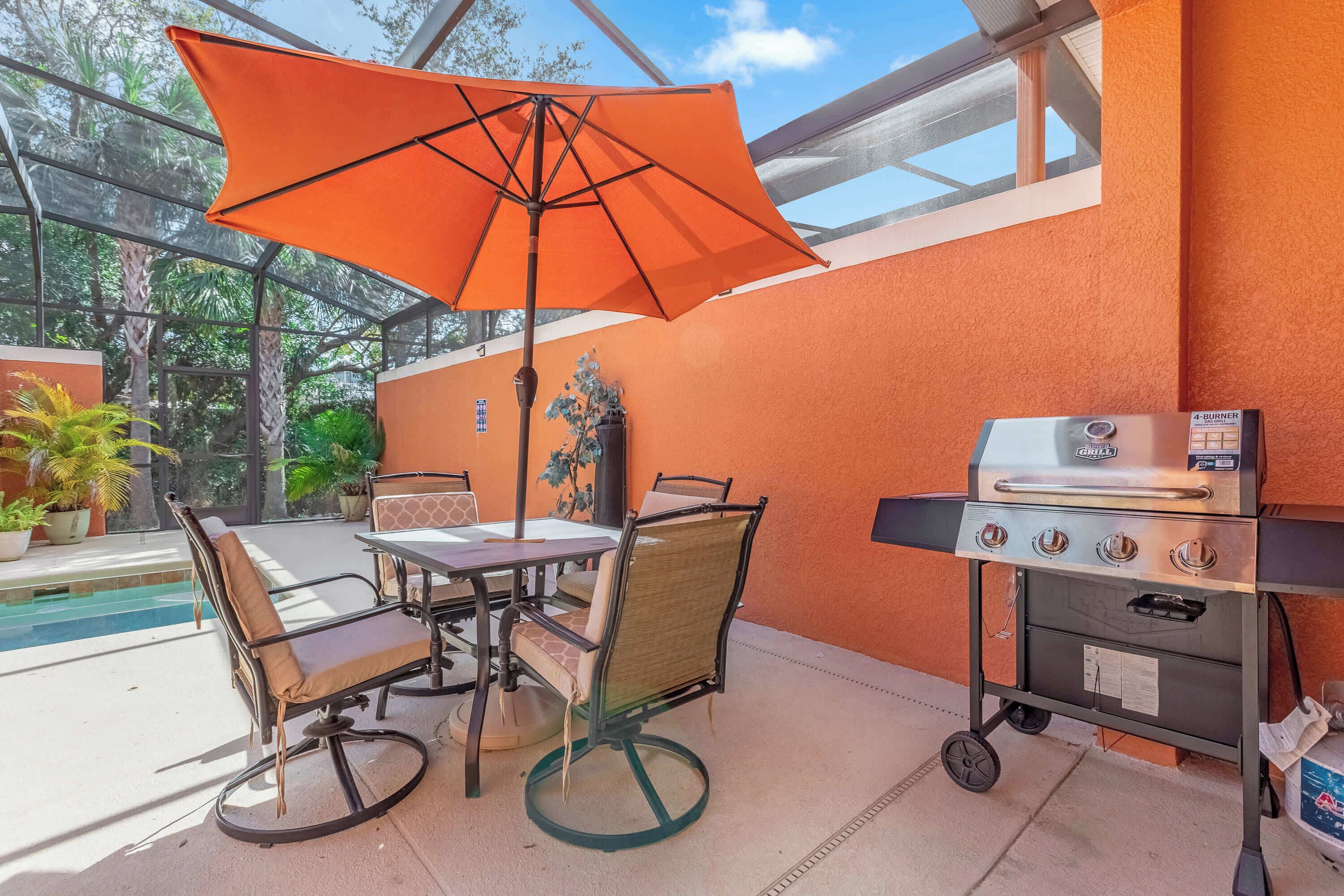 Un patio exterior con mesa y sillas, un paraguas naranja y una parrilla.