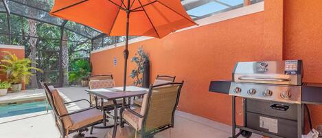 Un patio exterior con mesa y sillas, un paraguas naranja y una parrilla.