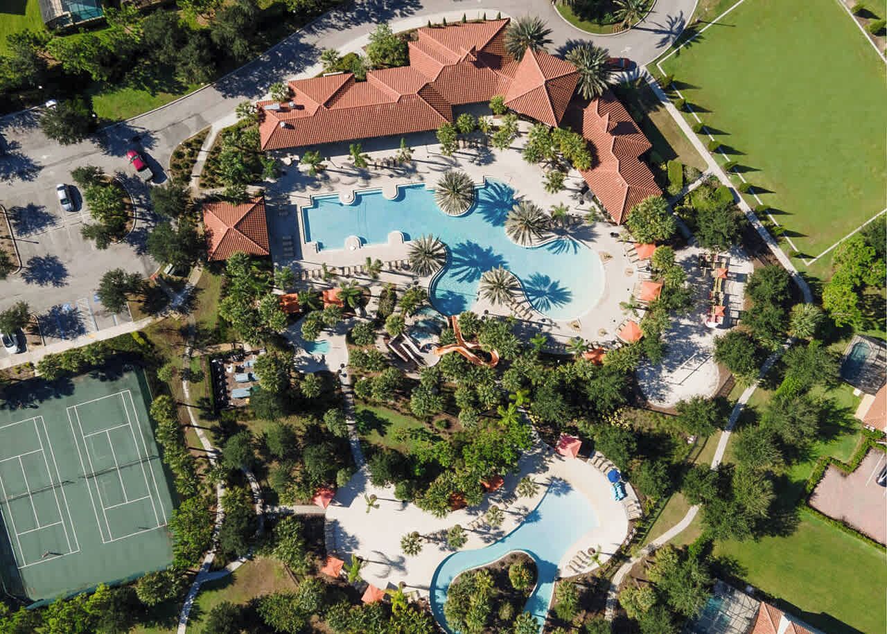 Oversigtsbillede af et resort med en central swimmingpool, tennisbaner og palmer.