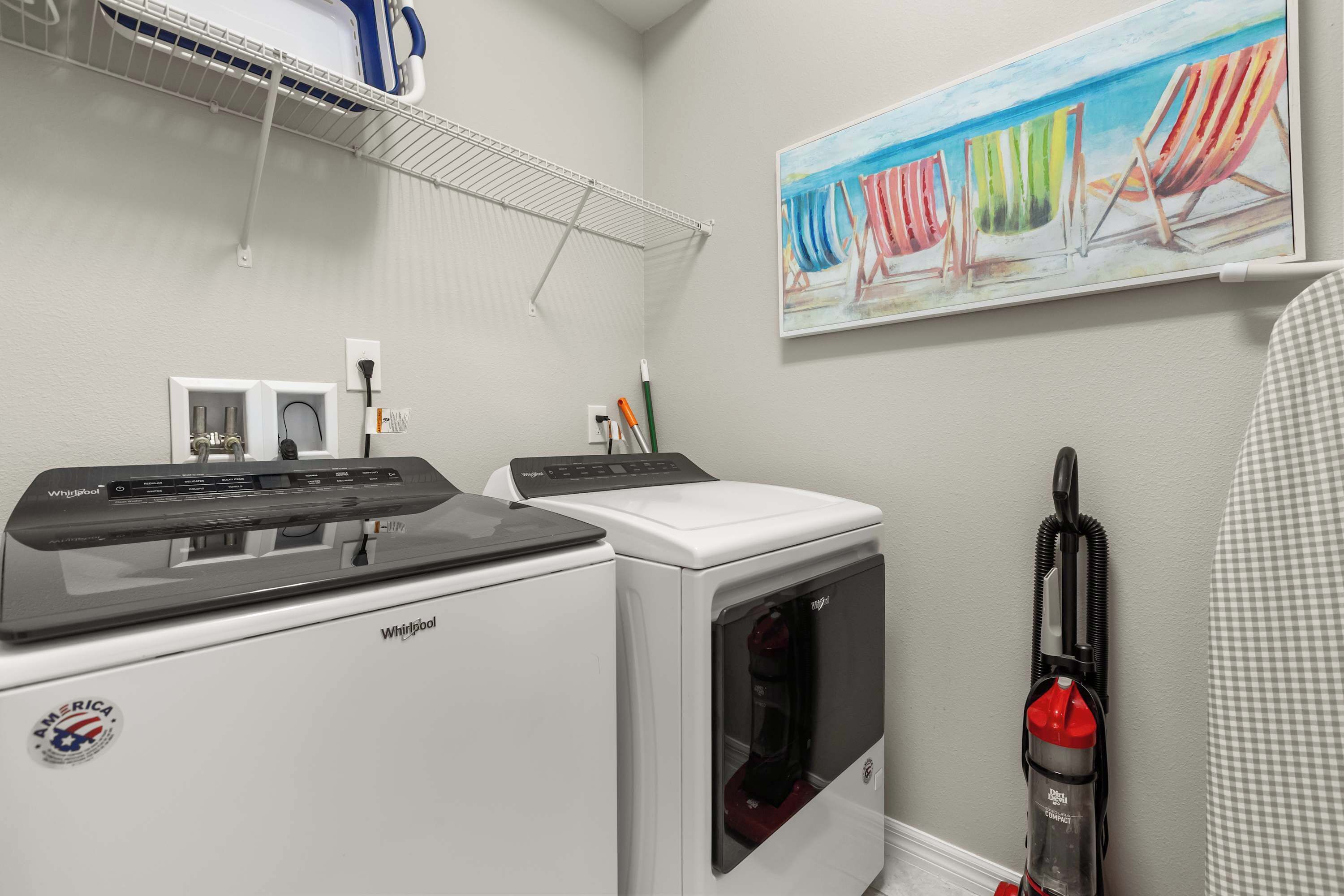 A laundry area with a Whirlpool washer and dryer, a wall-mounted shelf with a blue container, and a painting of beach chairs.