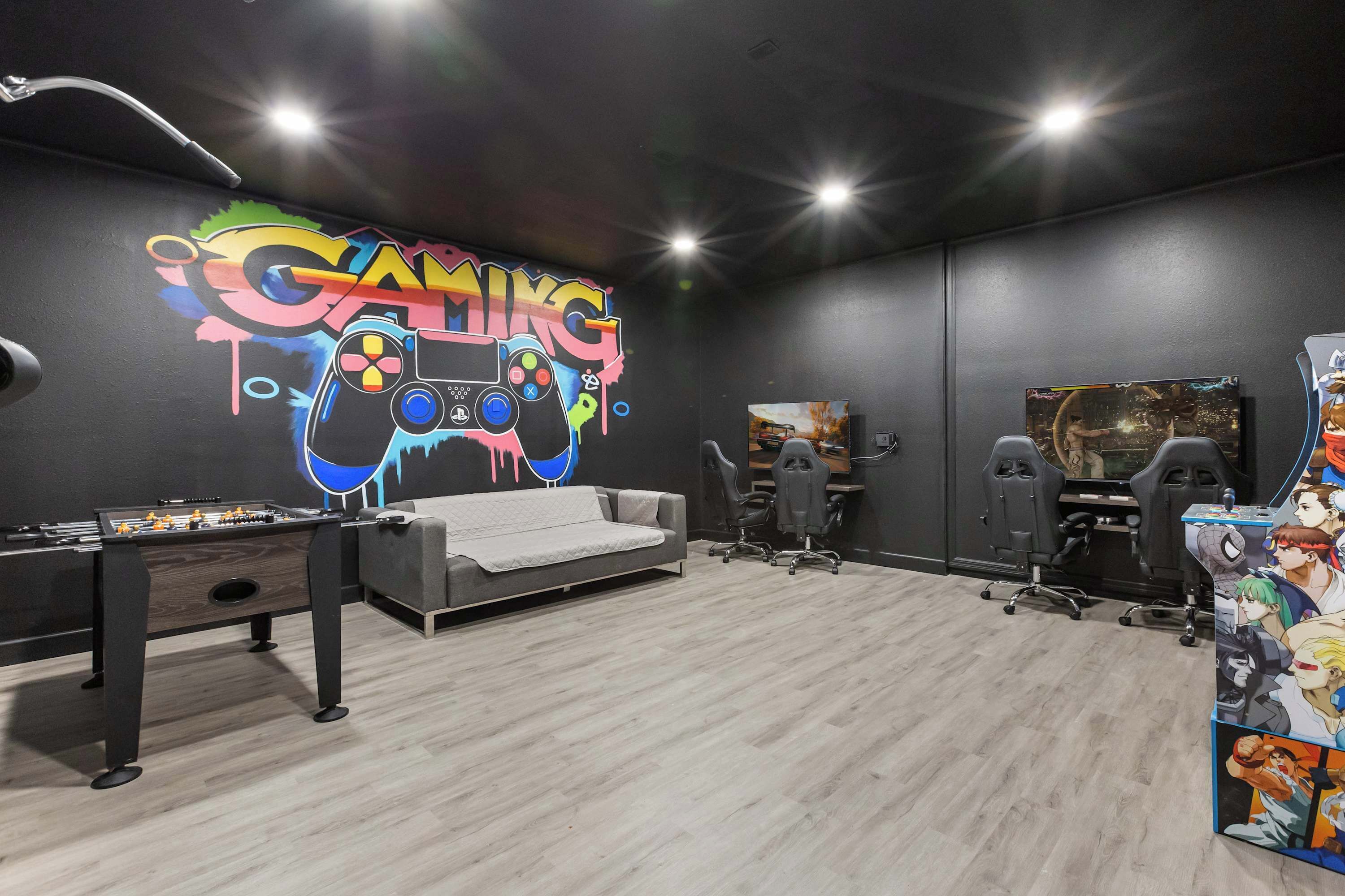 Una sala de juegos con mesa de fútbol, sillas gamer, un sofá y una pared con temática de videojuegos.