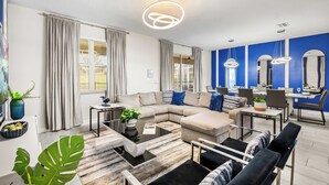 A modern living room with a sectional sofa, a coffee table, and a dining area with chairs and a table. - Upstay Villa Sleeps 26 - Theater, Arcade, Pool (Davenport)