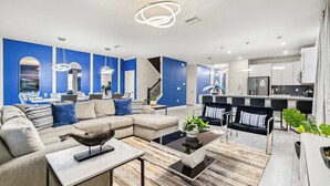 A modern living room with a sectional sofa, a coffee table, and a dining area with chairs. - Upstay Villa Sleeps 26 - Theater, Arcade, Pool (Davenport)