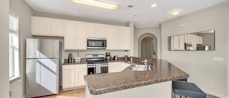 A modern kitchen with white cabinets, a stainless steel refrigerator, a microwave, and a granite countertop island with bar stools.