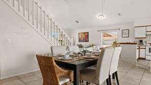 A dining area with a table set for four, surrounded by wicker chairs and white upholstered chairs. A staircase with white railings is visible to the left. - Upstay: Bright Family-friendly Retreat With Patio (Kissimmee)