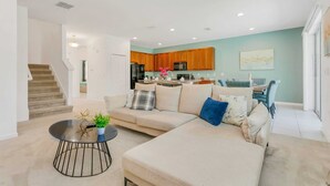 A modern living room with a sectional sofa, a round coffee table, and a kitchen in the background. - Resort Home w Bbq, Pool, Games- Near Disney! (Kissimmee)