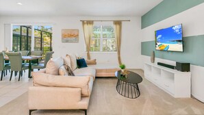 A modern living room with a beige sofa, a coffee table, and a flat-screen TV mounted on the wall. - Resort Home w Bbq, Pool, Games- Near Disney! (Kissimmee)