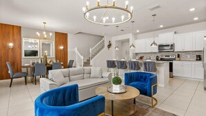 A modern living room with a sofa, two blue armchairs, a coffee table, and a dining area with a chandelier. - Upstay: Modern Windsor Resort Home Near Disney (Davenport)