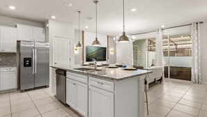 A modern kitchen with white cabinetry, a stainless steel refrigerator, a large island with a sink, pendant lighting, and a large window with a view of the outdoors. - Upstay: Modern Windsor Resort Home Near Disney (Davenport)