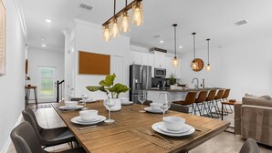 A modern dining area with a wooden table set for four, surrounded by black chairs. A kitchen with stainless steel appliances and a bar area is visible in the background. - Upstay: Luxury 4BR Family Home w Poolnear Disney (Davenport)
