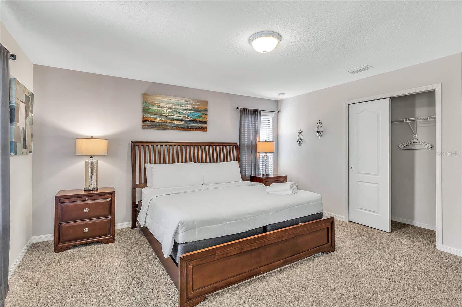 A bedroom with a wooden bed, two bedside tables with lamps, a painting on the wall, a window with curtains, and a closet with a hanger.