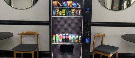 A vending machine with snacks and drinks, two wooden chairs, and a mirror on the wall.