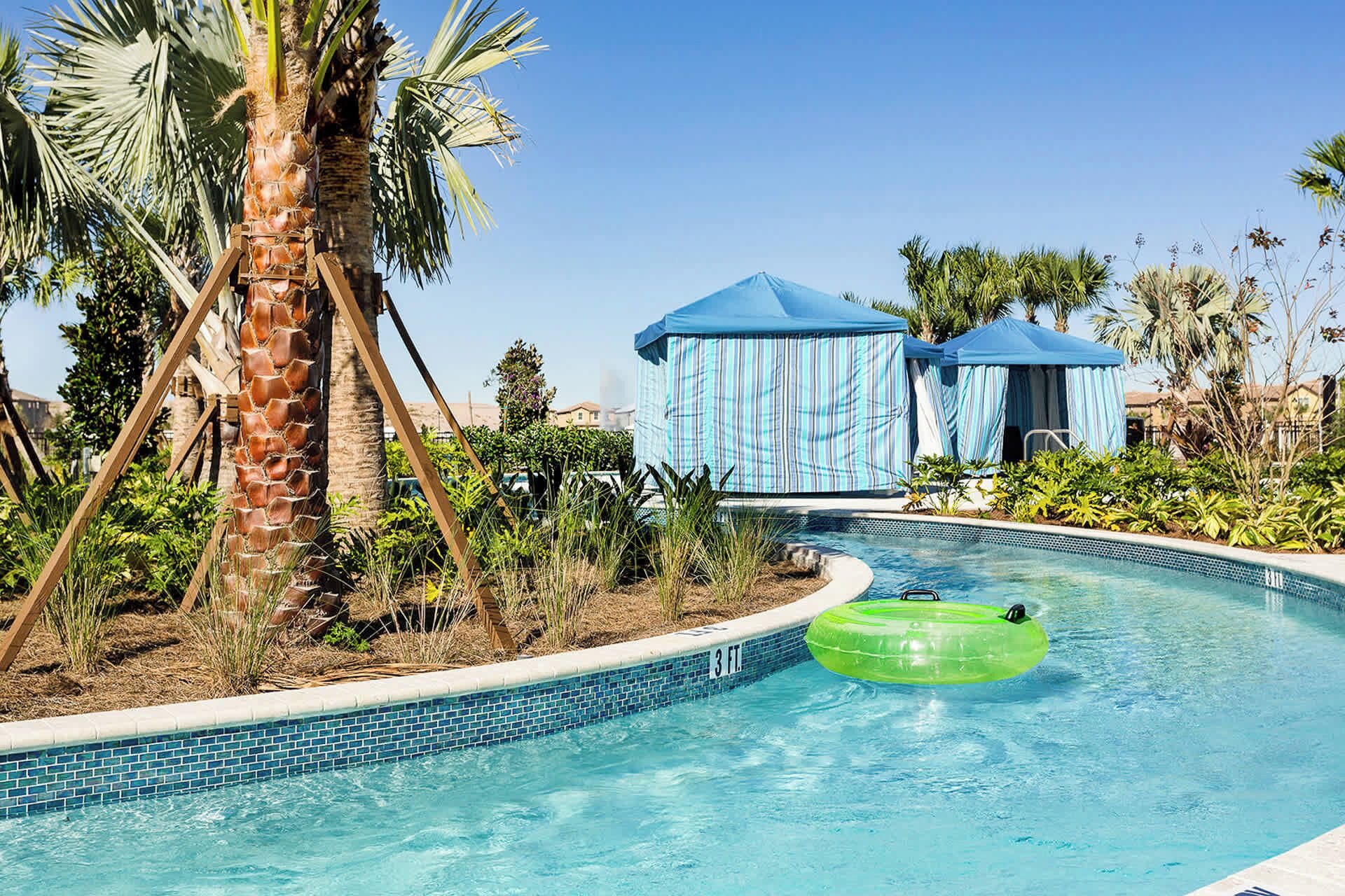 Una piscina con un flotador inflable verde, palmeras y toldos a rayas azules.
