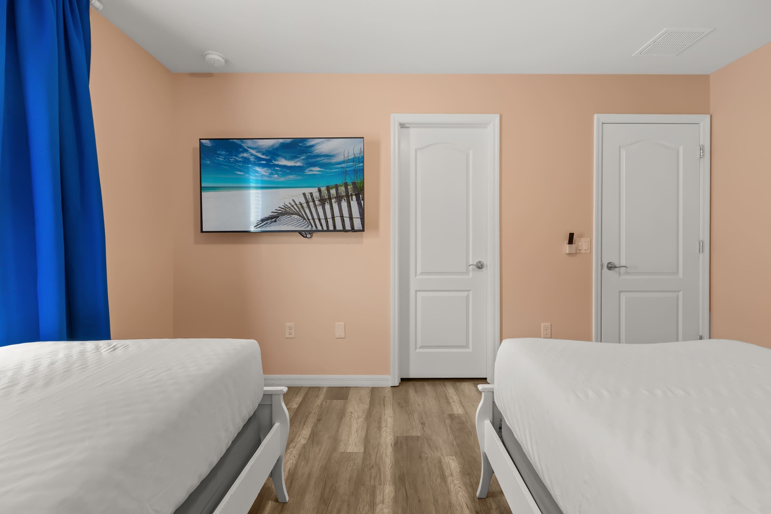 A bedroom with two beds, peach-colored walls, a painting of a beach scene, and white doors.