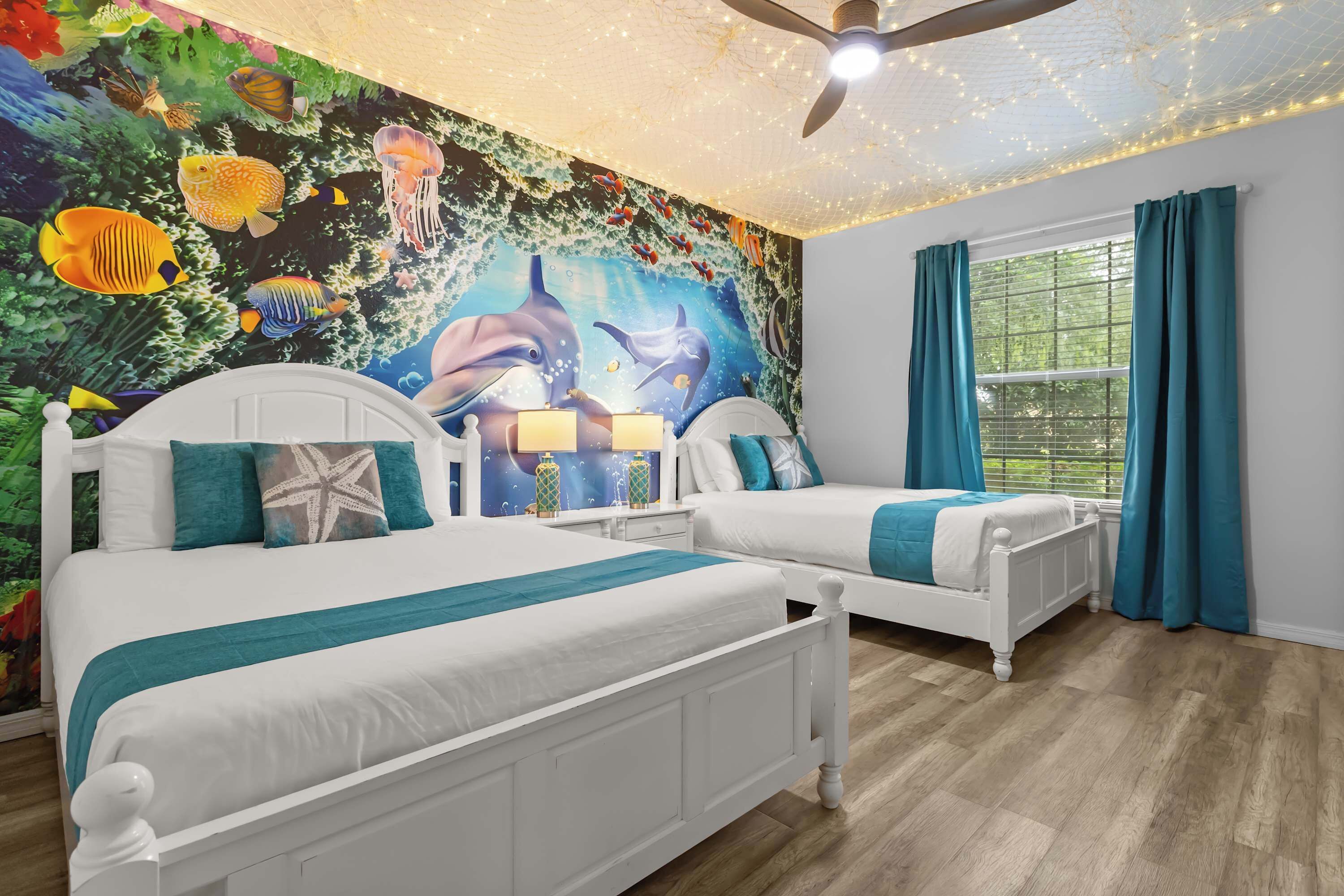 A bedroom with two beds, a mural of marine life, teal curtains, and a ceiling fan.