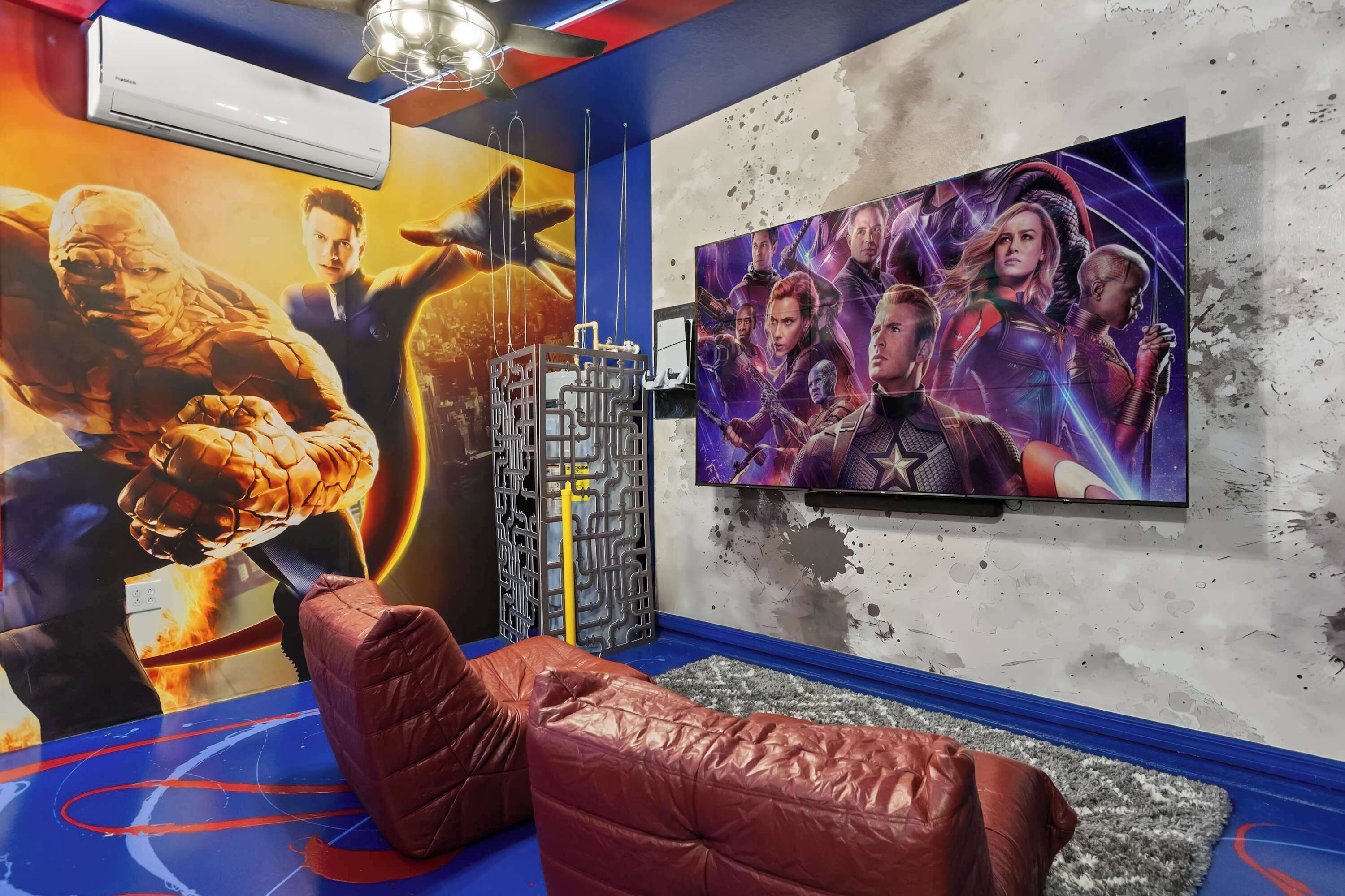 A room with a large TV on the wall, a red cushioned seating area, and a wall mural of a superhero movie.
