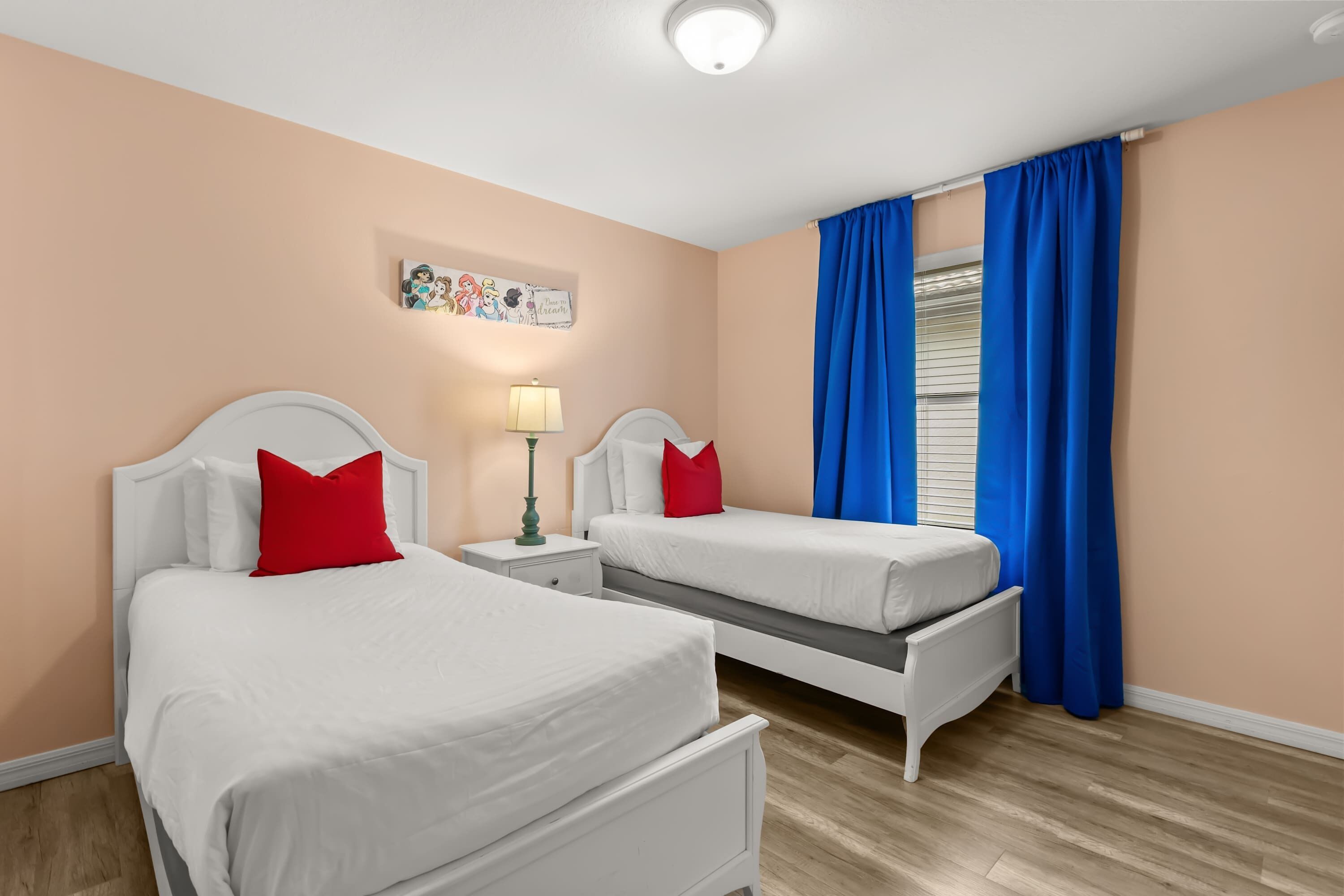 A hotel room with two single beds, a nightstand with a lamp, a wall decoration, and a window with blue curtains.