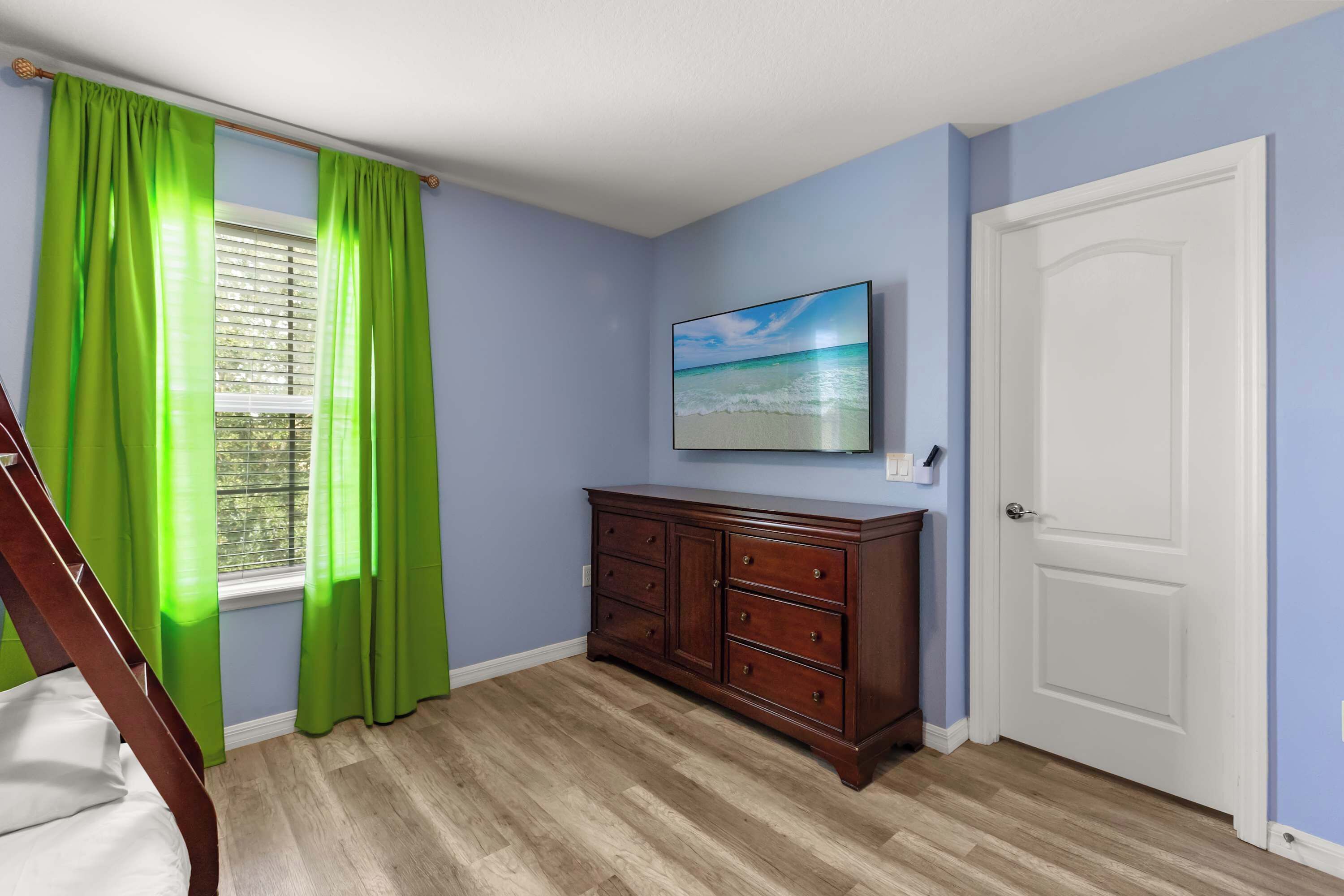 A room with a wooden dresser, a window with green curtains, a door, and a painting of a beach scene.
