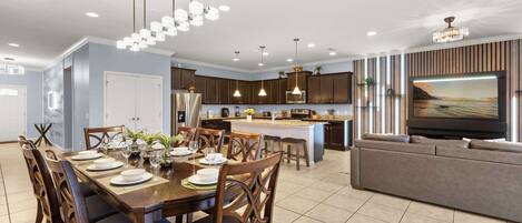 A modern kitchen with dark wooden cabinets, a central island, pendant lighting, and a large flat-screen TV mounted on the wall.