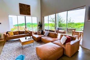 A spacious living room with large windows, a brown leather sofa set, a coffee table, and a view of greenery. - Luxury Hilltop Villa With Hot Tub & Scenic Views (Dripping Springs)