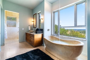 A bathroom with a freestanding bathtub, a wooden vanity, a large mirror, and a view of the outdoors. - Luxury Hilltop Retreat w/ Scenic Views (Dripping Springs)