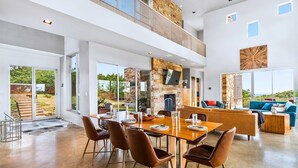 A spacious living area with a high ceiling, a stone fireplace, and a large dining table set for six. - Luxury Hilltop Villa With Hot Tub & Scenic Views (Dripping Springs)