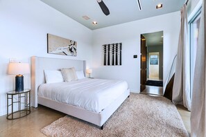 A modern bedroom with a large bed, two bedside lamps, a wall-mounted artwork, a decorative curtain, and a hallway leading to another room. - Luxury Hilltop Villa With Hot Tub & Scenic Views (Dripping Springs)