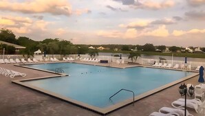 A rectangular swimming pool with multiple lanes, surrounded by white lounge chairs and umbrellas, with a view of a lake and trees in the background. - Upstay 2BR Golfer's Retreat w Patio Ventura Resort (Orlando)
