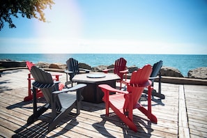 A wooden deck with Adirondack chairs and a round table, overlooking a calm sea. - On The Rocks Lake House – Your Gateway to Point Pelee National Park Adventures (Leamington)