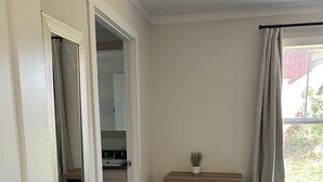Een kamer met een houten vloer, een witte deur met spiegel, een klein houten plankje met manden, en een raam met gordijnen.
