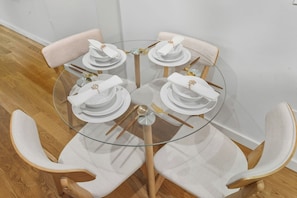 A round glass table with four white plates, each with a folded napkin and a gold napkin ring. The table is surrounded by four wooden chairs with white cushions. - Elegant Studio Apartment in the Heart of Astoria (ASTORIA)