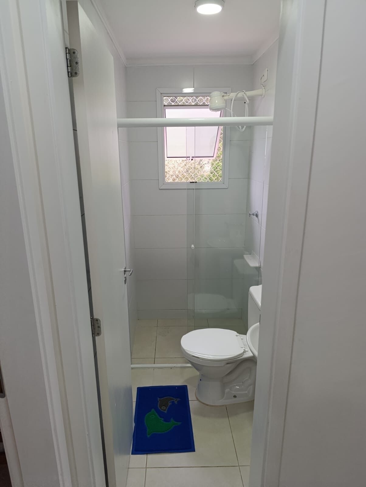 Une petite salle de bain blanche, équipée de toilettes, d’une douche et d’une fenêtre ornée d’une grille décorative.