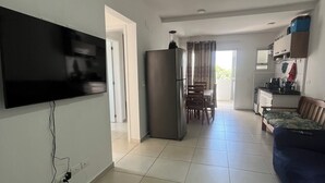 A spacious living area with a flat-screen TV mounted on the wall, a large refrigerator, a dining table with chairs, a sofa, and a kitchen area with cabinets and a countertop. - Beautiful, cozy apartment in Pereque Açu, Passaredo, Ubatuba. (Ubatuba)