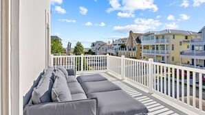 Een balkon met grijs gestoffeerde zitplaatsen, witte relingen en uitzicht op kleurrijke gebouwen en een heldere lucht.